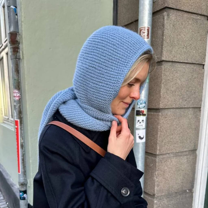 Frau mit blauem Sophie Hood von PetiteKnit steht im Mantel vor einem Geschäft.