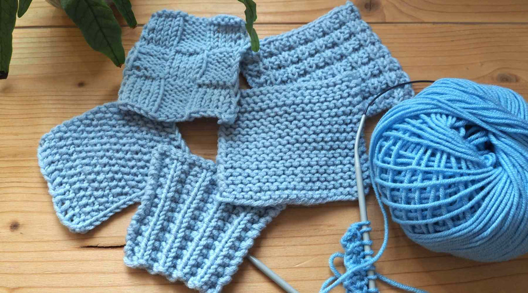 Fünf Maschenproben aus hellblauem Garn mit einfachen Strickmustern auf einem Holztisch, neben einem hellbauen Wollknäuel.