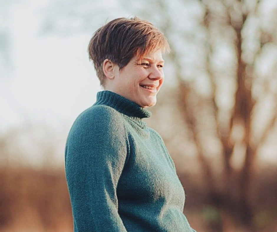 Smiling person in a green sweater, outdoors with trees in the background.