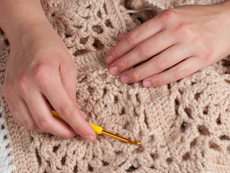 Eine Grannysquaredecke mit zwei Händen und einer goldenen Häkelnadel.