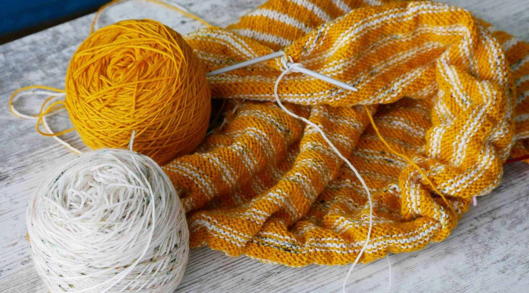 Unfertiges Sommershirt mit gelben und weißen Streifen auf Stricknadeln.